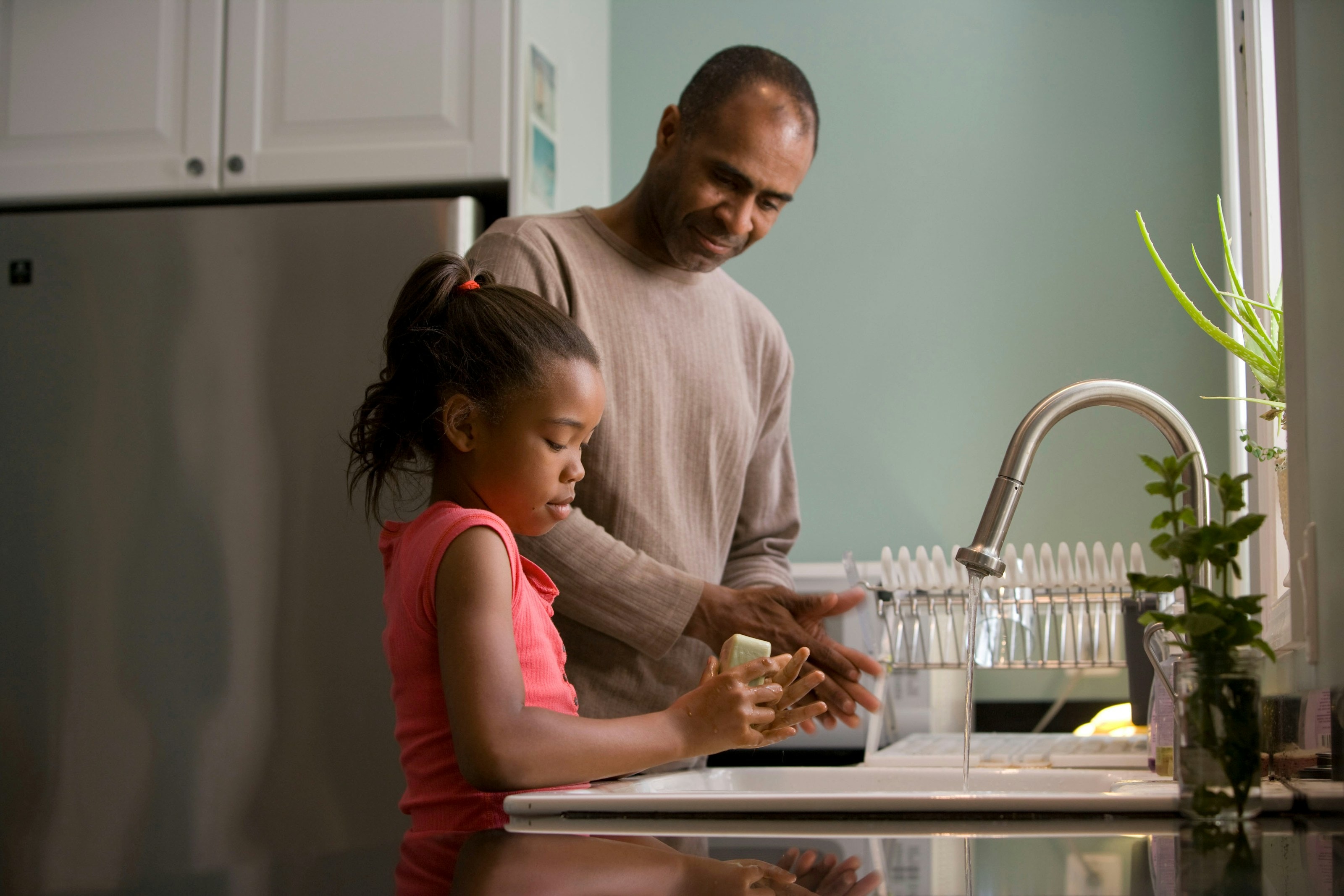 A father and his daughter washing up together at a kitchen sink in daylight.