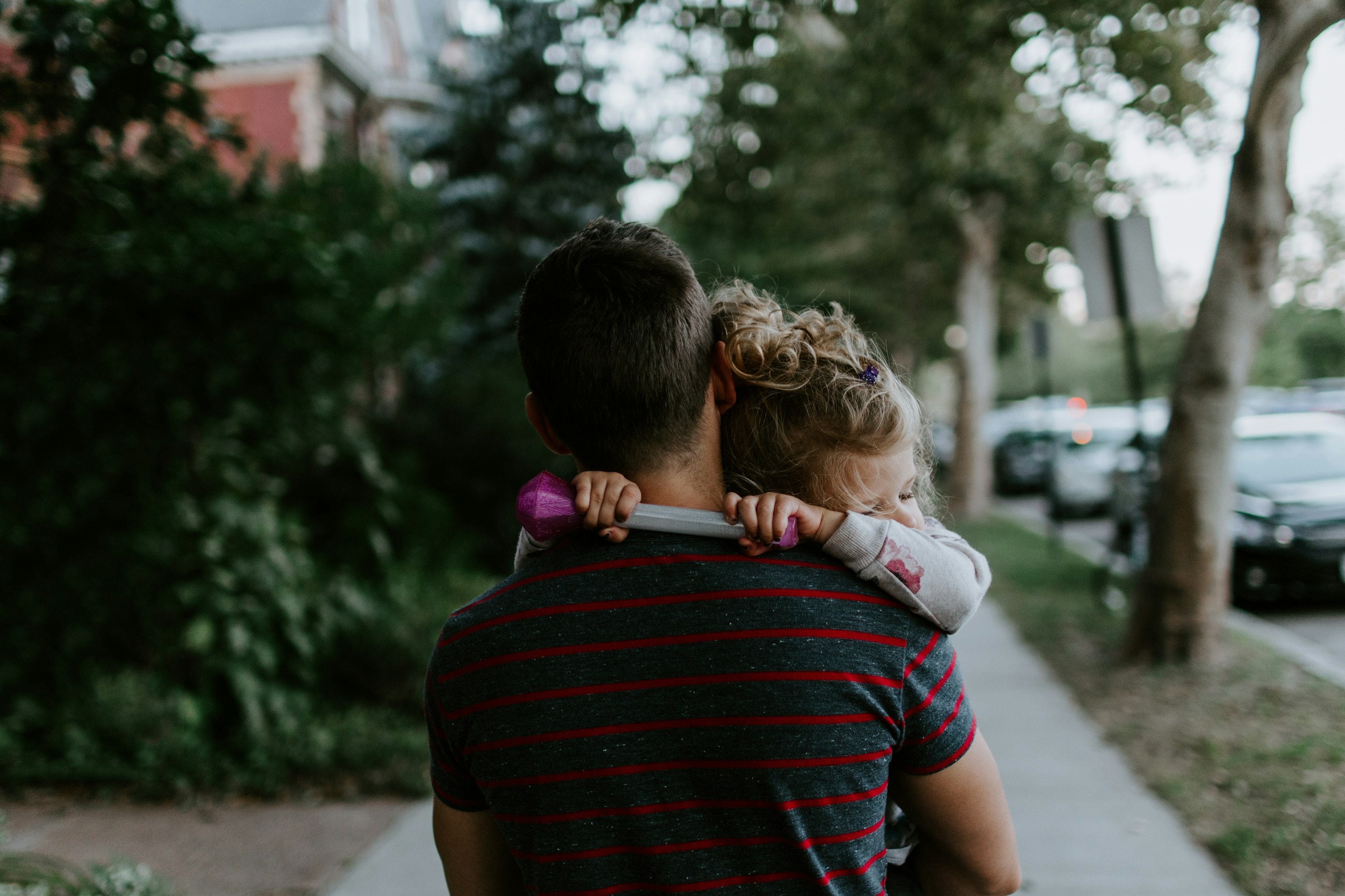 A parent carrying a tired child home along a tree-lined street.